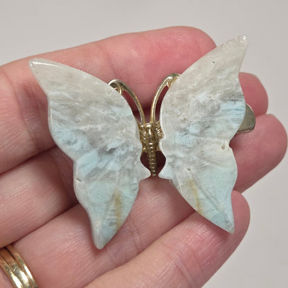 A butterfly-shaped brooch with iridescent pale blue and white wings and gold-tone metal hardware, shown resting on a person's fingers.