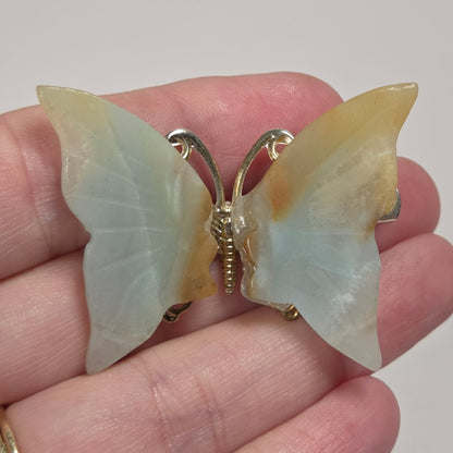 A butterfly-shaped brooch with iridescent pale blue-green and yellow wings, held on a person's hand. The brooch features a gold-tone metal butterfly body and antennae.