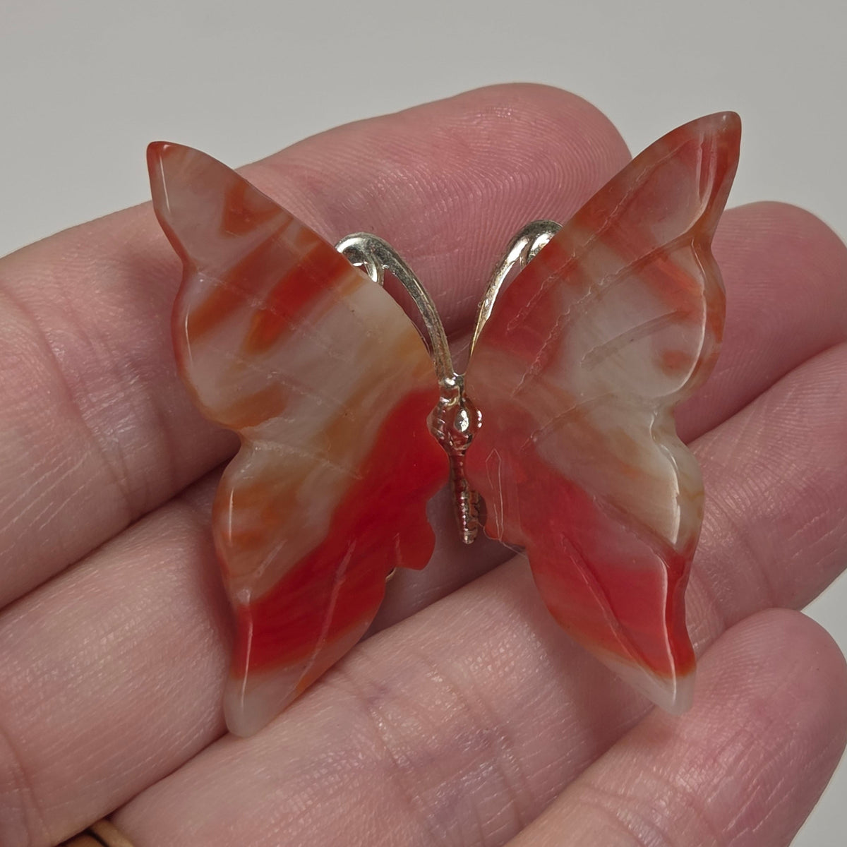A butterfly-shaped brooch made from red and cream banded agate, held in a person's hand. The butterfly has a silver-tone metal wire frame and a small gold-tone ring at the center.