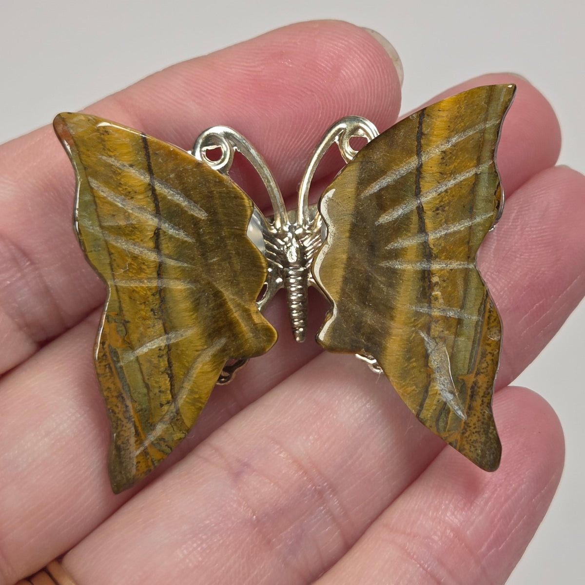 A butterfly-shaped brooch with textured golden-brown wings and a silver metal body, held on a person's fingers against a light gray background.