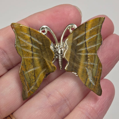 A butterfly-shaped brooch with textured golden-brown wings and a silver metal body, held on a person's fingers against a light gray background.