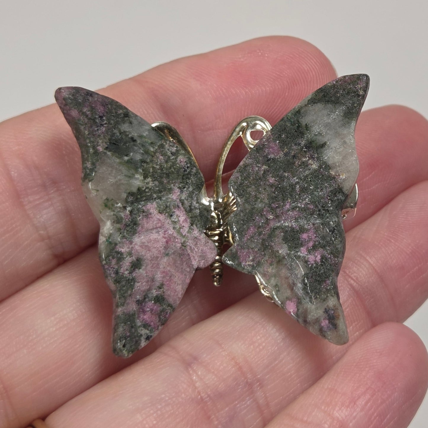 A butterfly-shaped brooch with mottled green, pink, and gray stone wings and a gold-tone metal back, held on a person's fingers against a plain light background.