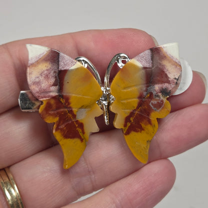 A butterfly-shaped brooch with yellow and brown marbled wings and silver-tone metal details, held in a person's hand against a plain light background.