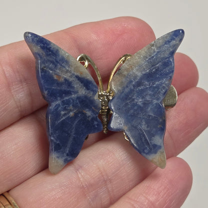 A butterfly-shaped brooch made of blue lapis lazuli stone with gold-tone metal accents, held on a person's hand.