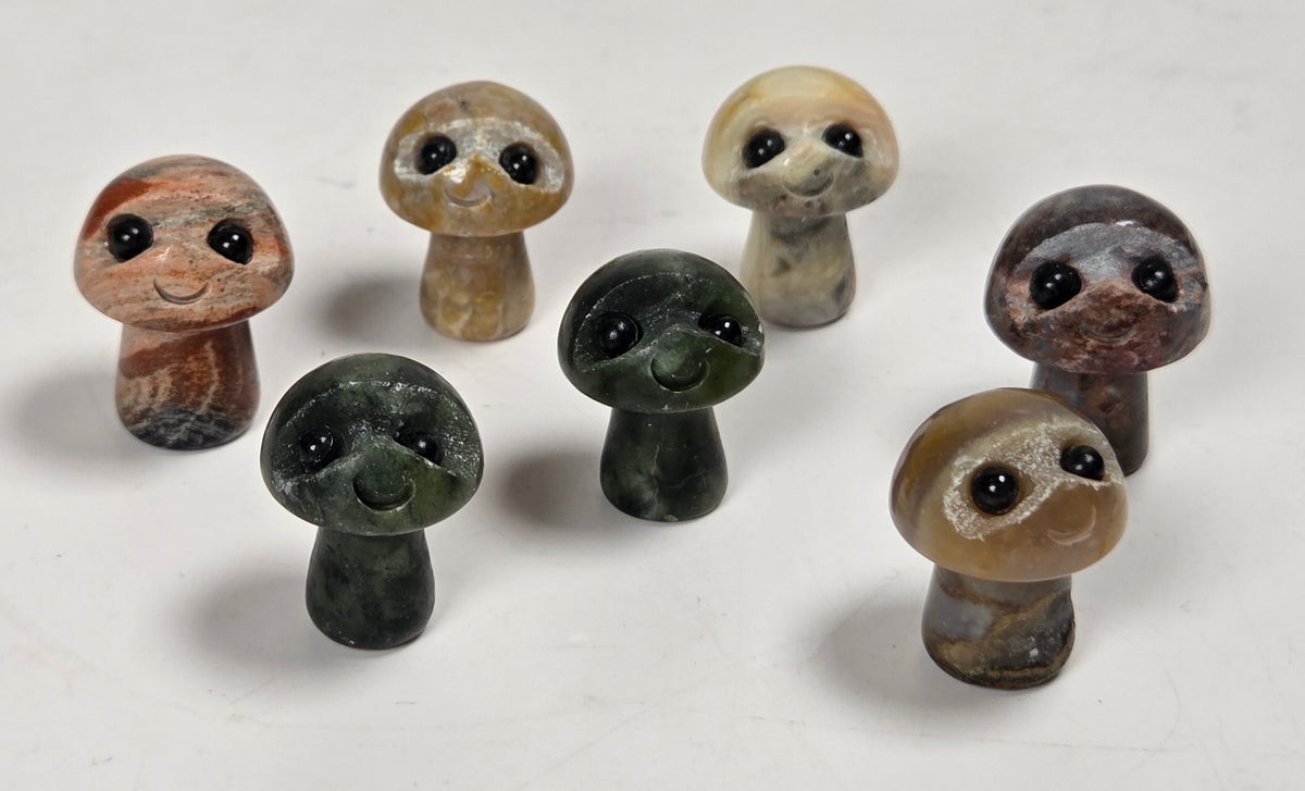 Seven small carved stone mushroom figurines with cute cartoon faces, displayed on a plain off-white surface.