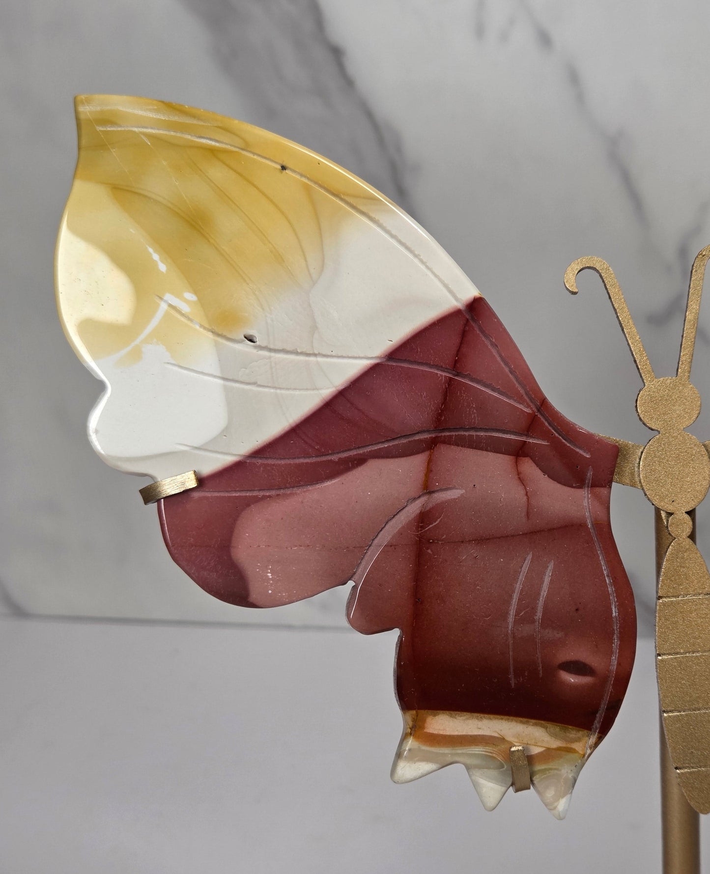 mookaite crystal carved butterfly wings on a gold stand against a marble background