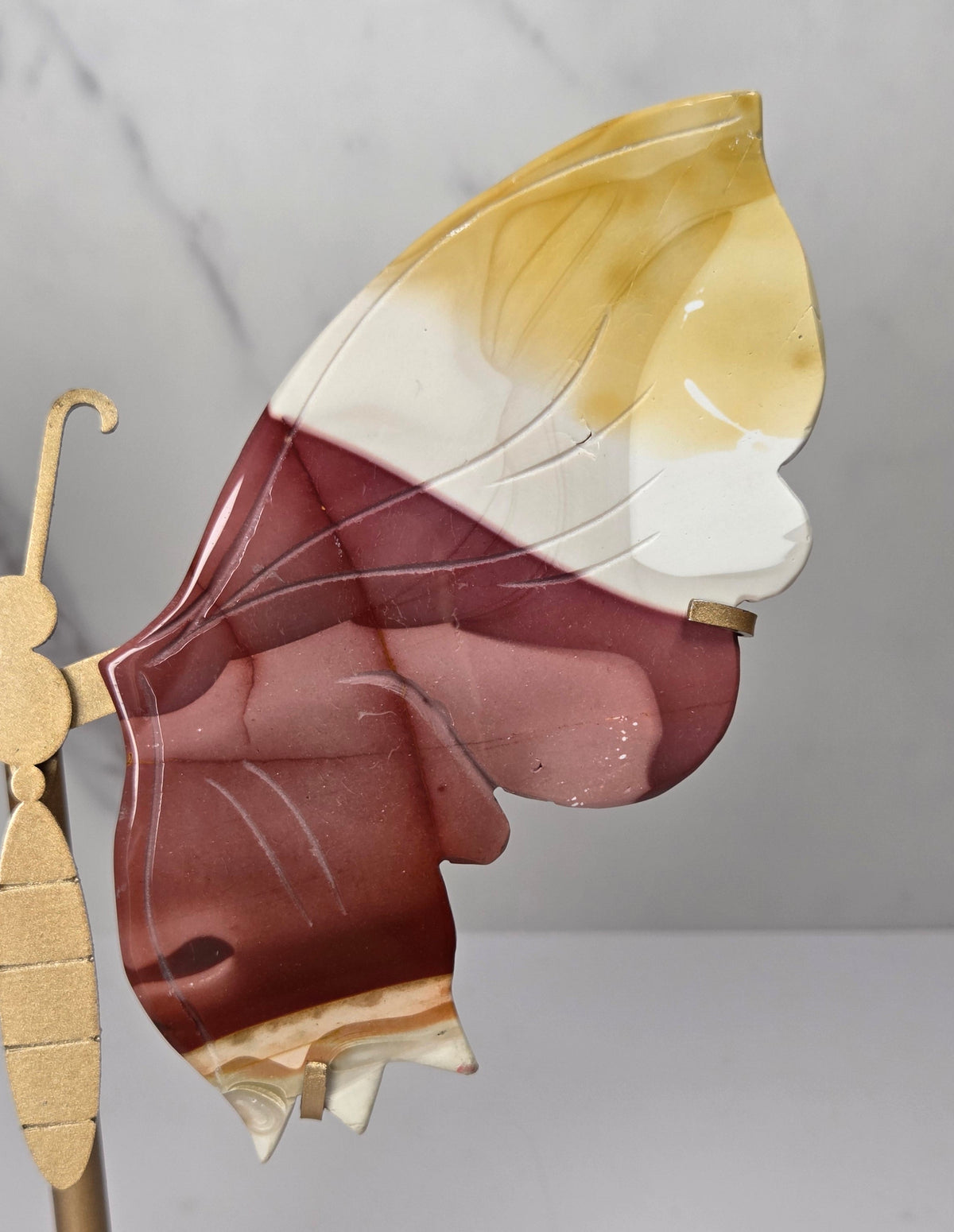 mookaite crystal carved butterfly wings on a gold stand against a marble background