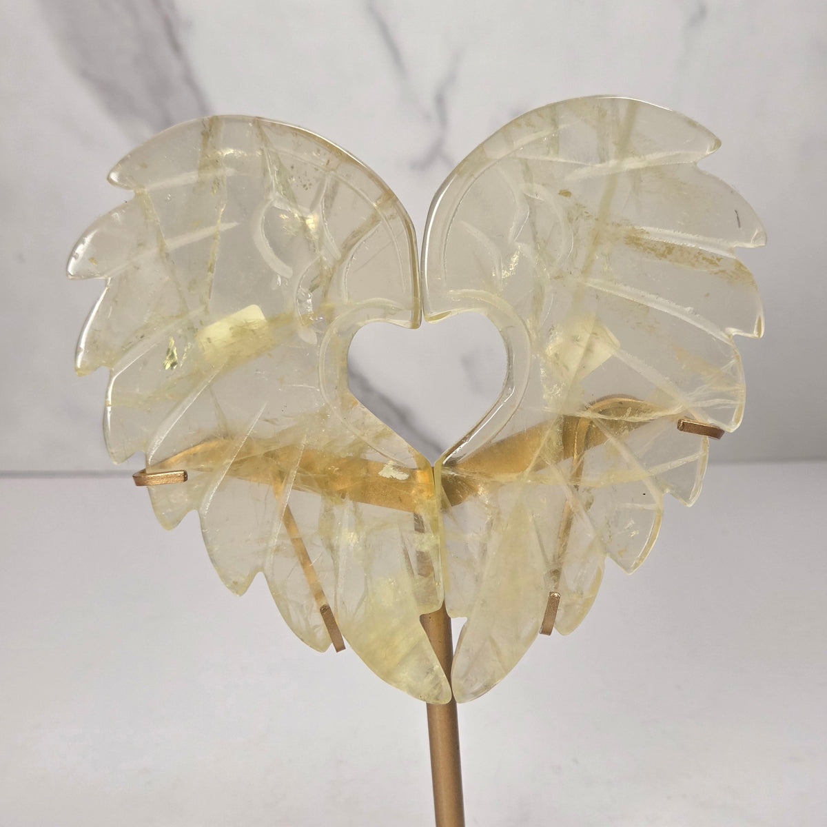 Carved Citrine wings that are in the shape of a heart, on a gold stand against a marbled background