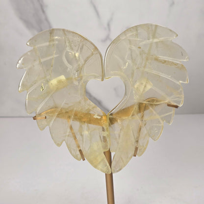 Carved Citrine wings that are in the shape of a heart, on a gold stand against a marbled background