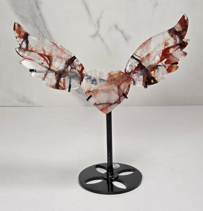 A decorative metal angel wing sculpture with a polished heart-shaped center, mounted on a black stand against a white marble-patterned background.
