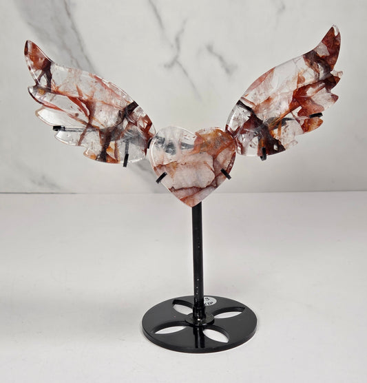 A decorative metal angel wing sculpture with a polished heart-shaped center, mounted on a black stand against a white marble-patterned background.