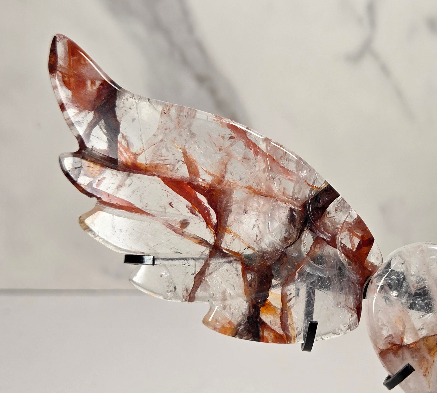 A decorative metal angel wing sculpture with a polished heart-shaped center, mounted on a black stand against a white marble-patterned background.