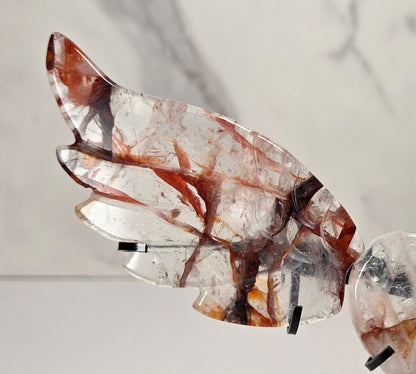 A decorative metal angel wing sculpture with a polished heart-shaped center, mounted on a black stand against a white marble-patterned background.