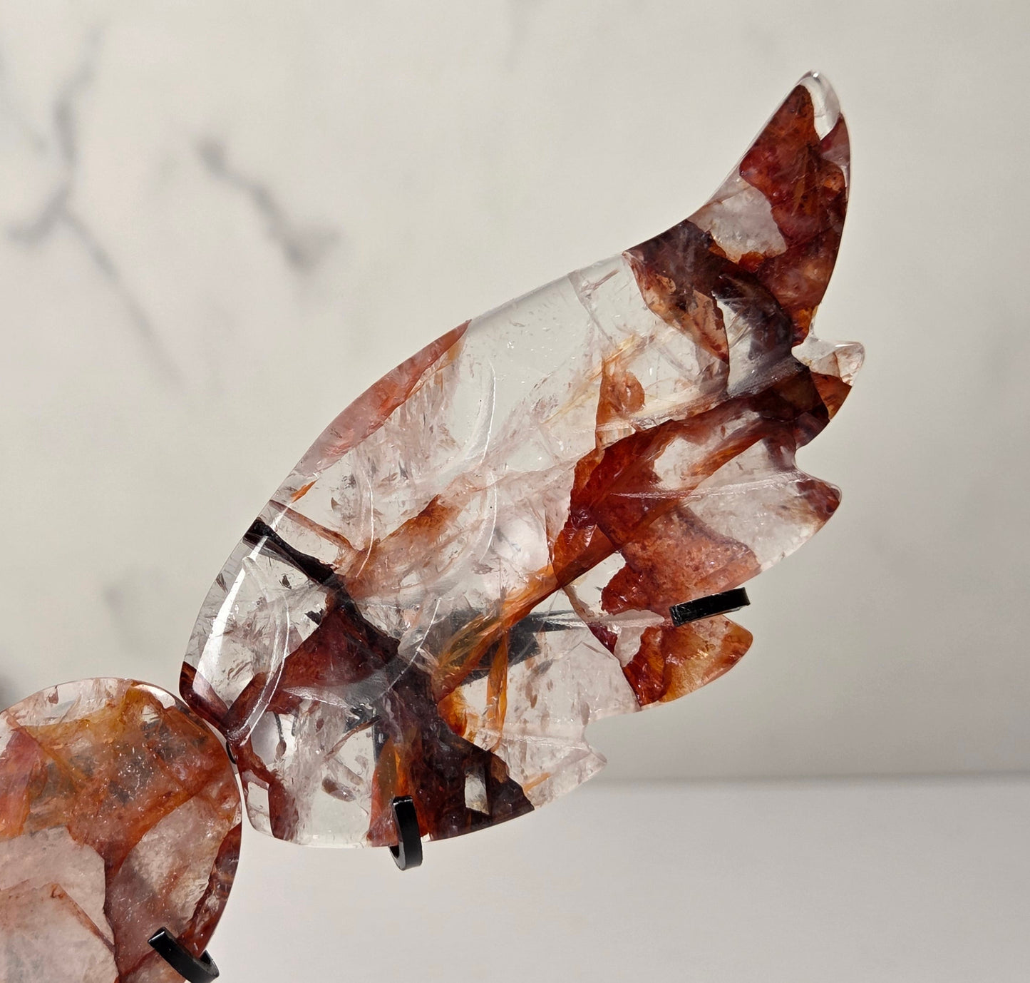 A decorative metal angel wing sculpture with a polished heart-shaped center, mounted on a black stand against a white marble-patterned background.