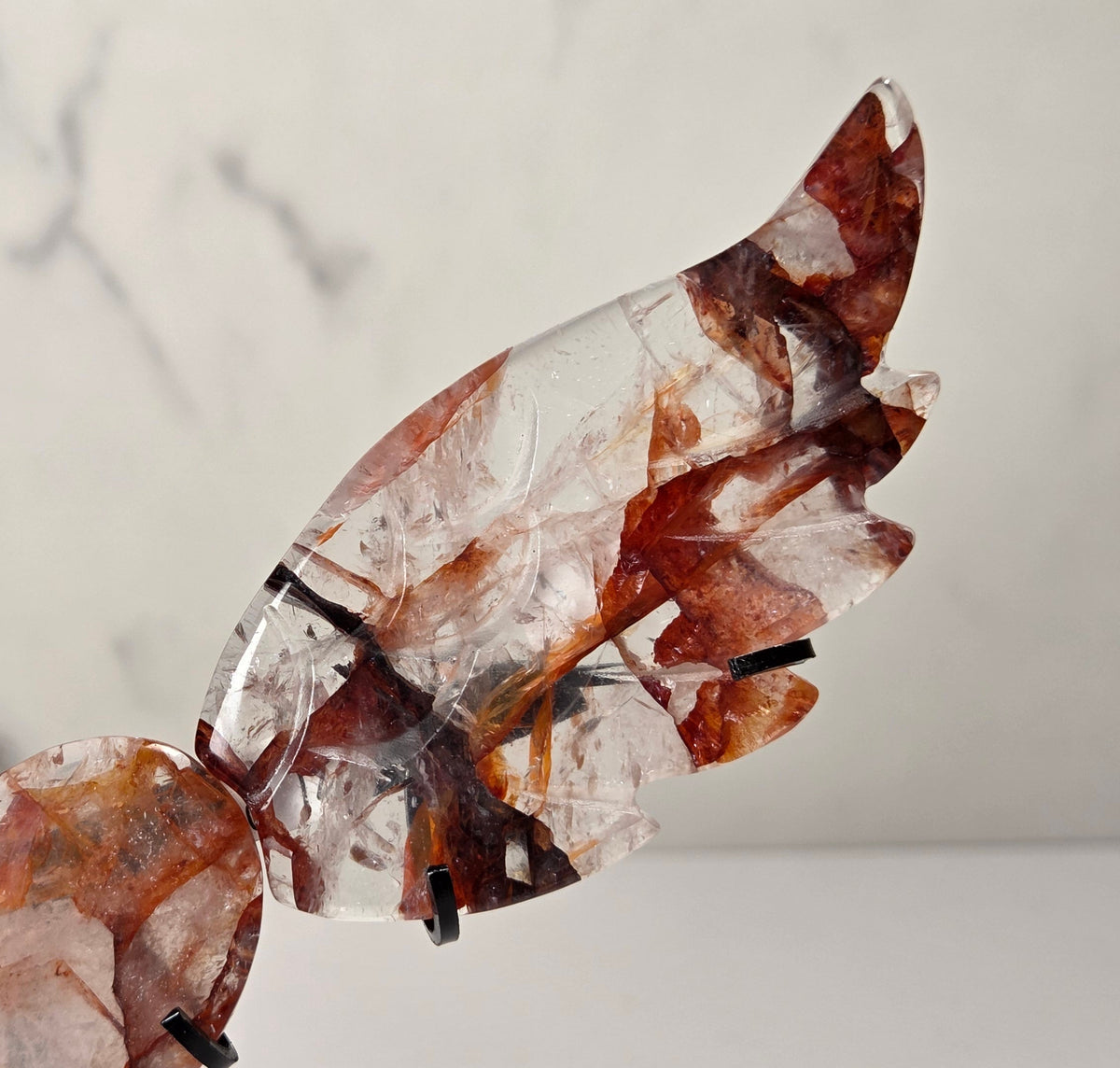 A decorative metal angel wing sculpture with a polished heart-shaped center, mounted on a black stand against a white marble-patterned background.