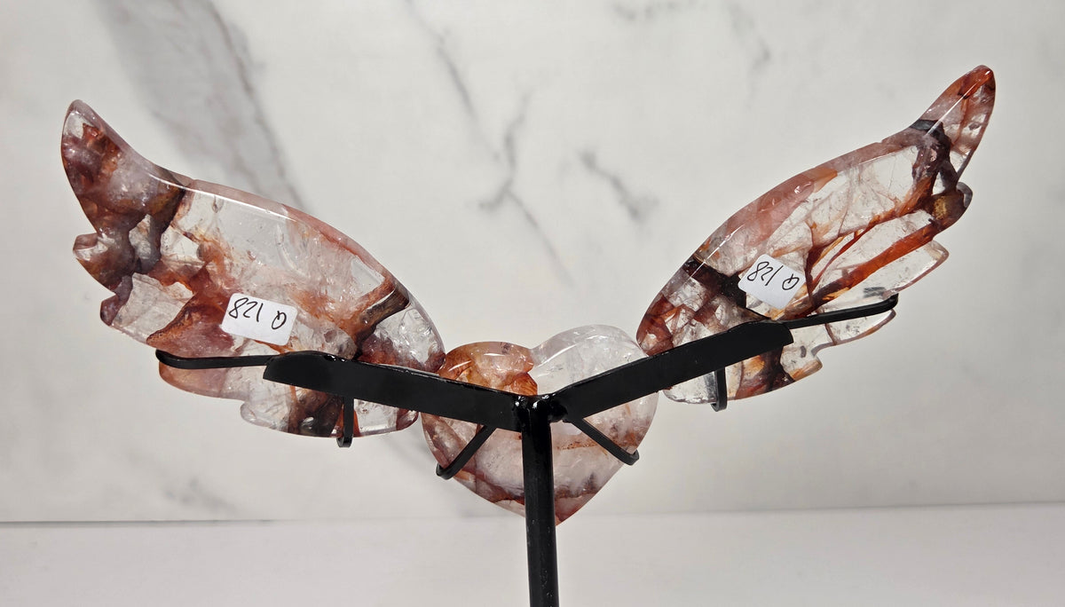 A decorative metal angel wing sculpture with a polished heart-shaped center, mounted on a black stand against a white marble-patterned background.