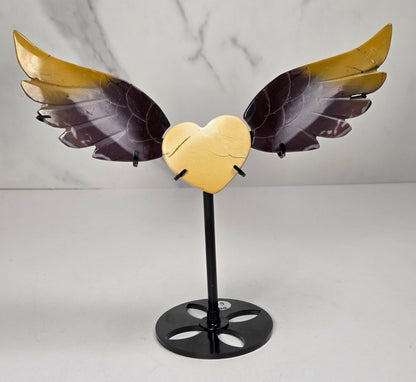 A decorative metal angel wing sculpture with a gold heart at the center, mounted on a black stand against a light gray marble-patterned background.