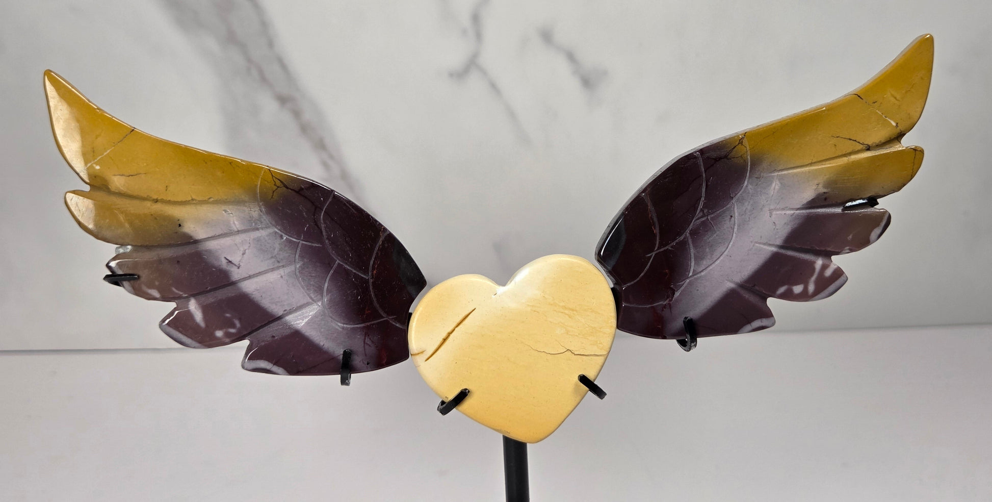 A decorative metal sculpture of a cracked gold heart with two wings, one wing gold and the other dark brown, mounted on a black stand against a light gray marble-patterned background.