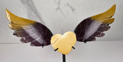 A decorative metal sculpture of a cracked gold heart with two wings, one wing gold and the other dark brown, mounted on a black stand against a light gray marble-patterned background.