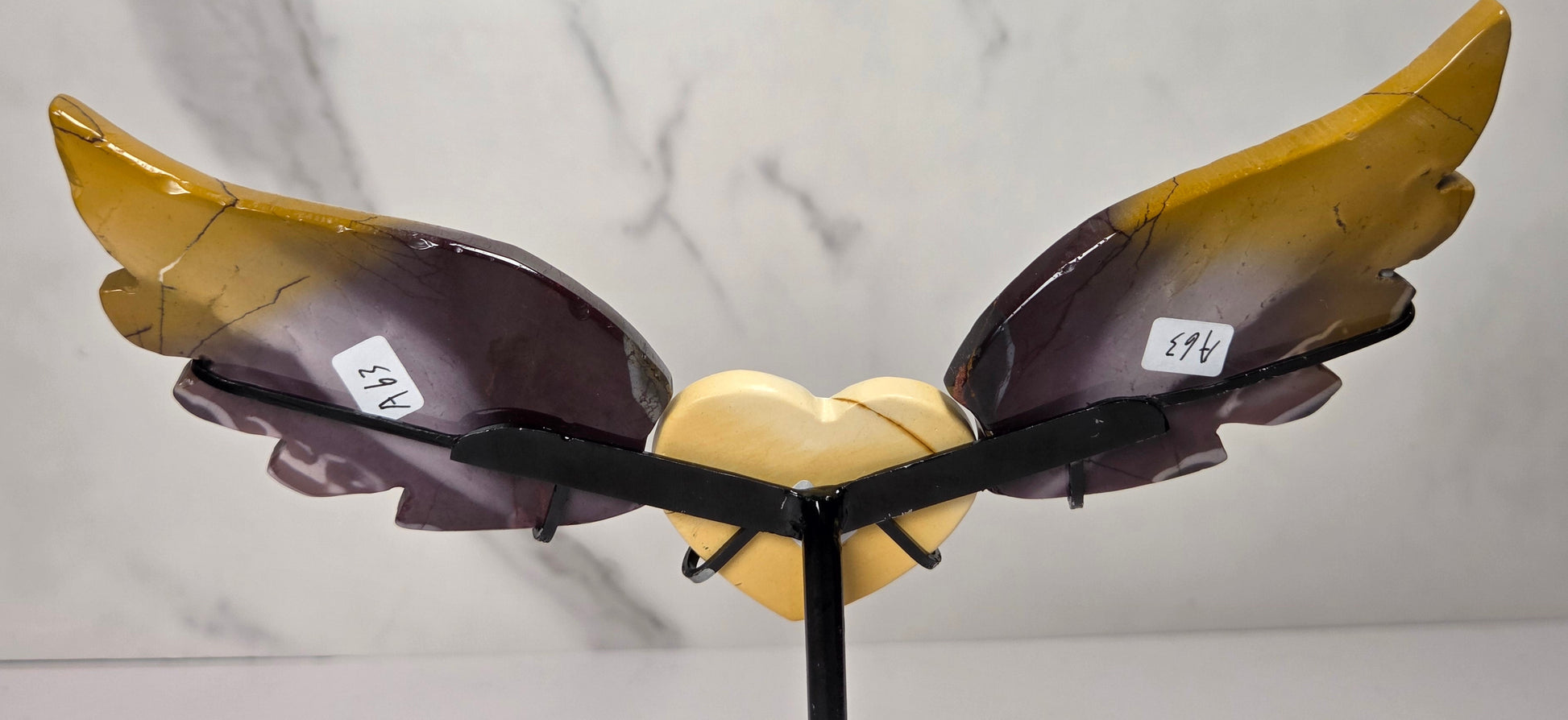 A decorative winged heart sculpture with a glossy amber-yellow and dark purple gradient finish, mounted on a black stand against a light gray marble-patterned background.