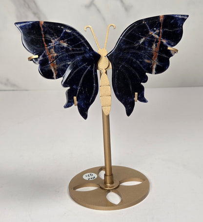 A butterfly-shaped jewelry display stand made of deep blue lapis lazuli stone with gold-toned metal accents, shown on a light marble background.
