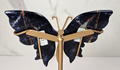 A butterfly-shaped jewelry display stand made of polished dark blue lapis lazuli stone with gold-tone metal arms and stand, shown against a light marble-patterned background.