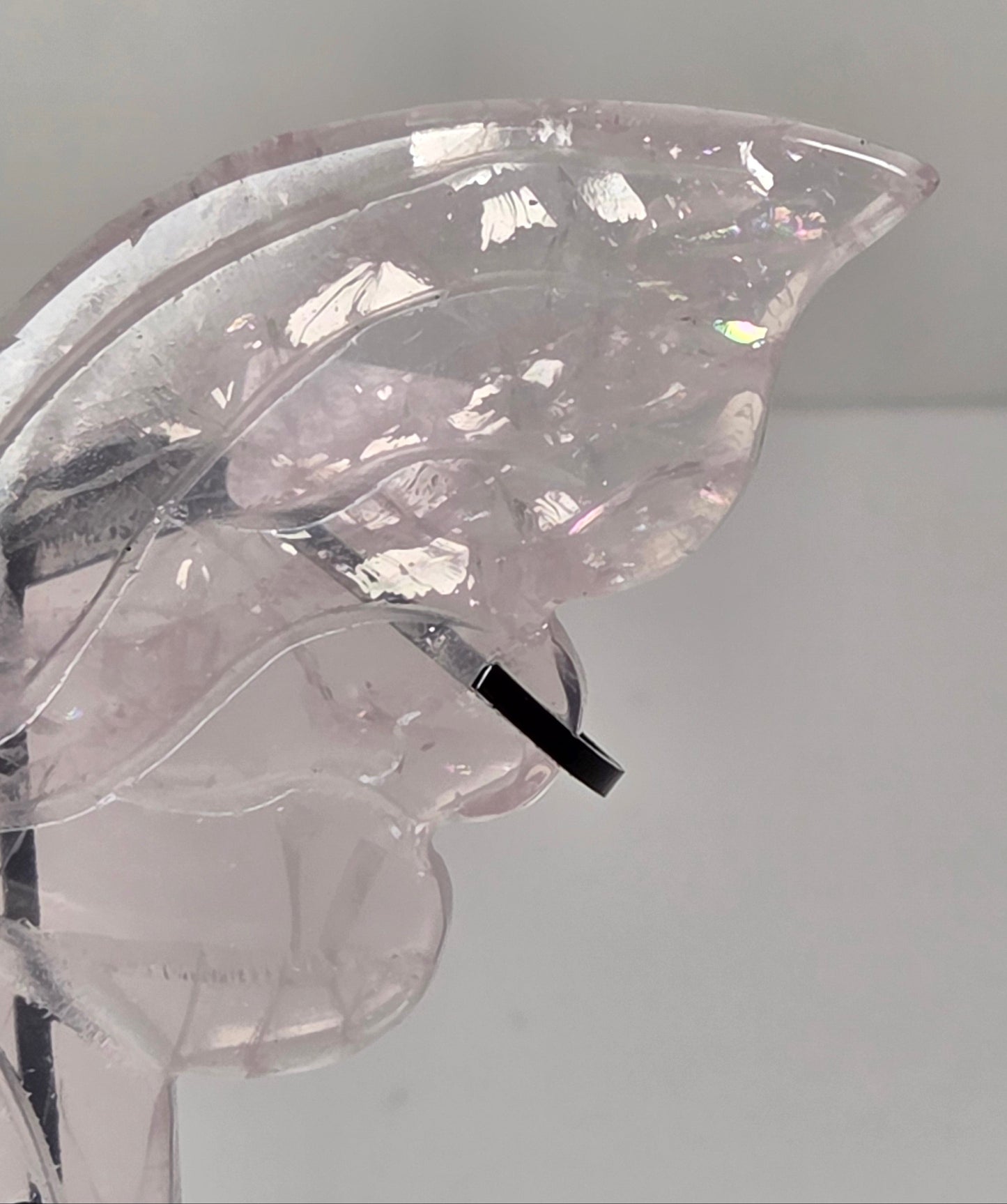 A close-up of a translucent pale pink crystal or glass object with a curved, organic shape and a small black rectangular accent, photographed against a plain light background.