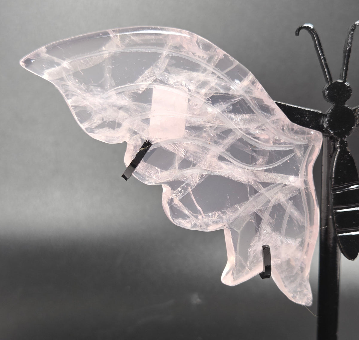 A butterfly-shaped wall hanging made from translucent pale pink stone with black metal supports, photographed against a dark gray studio background.