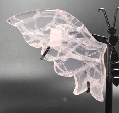 A butterfly-shaped wall hanging made from translucent pale pink stone with black metal supports, photographed against a dark gray studio background.