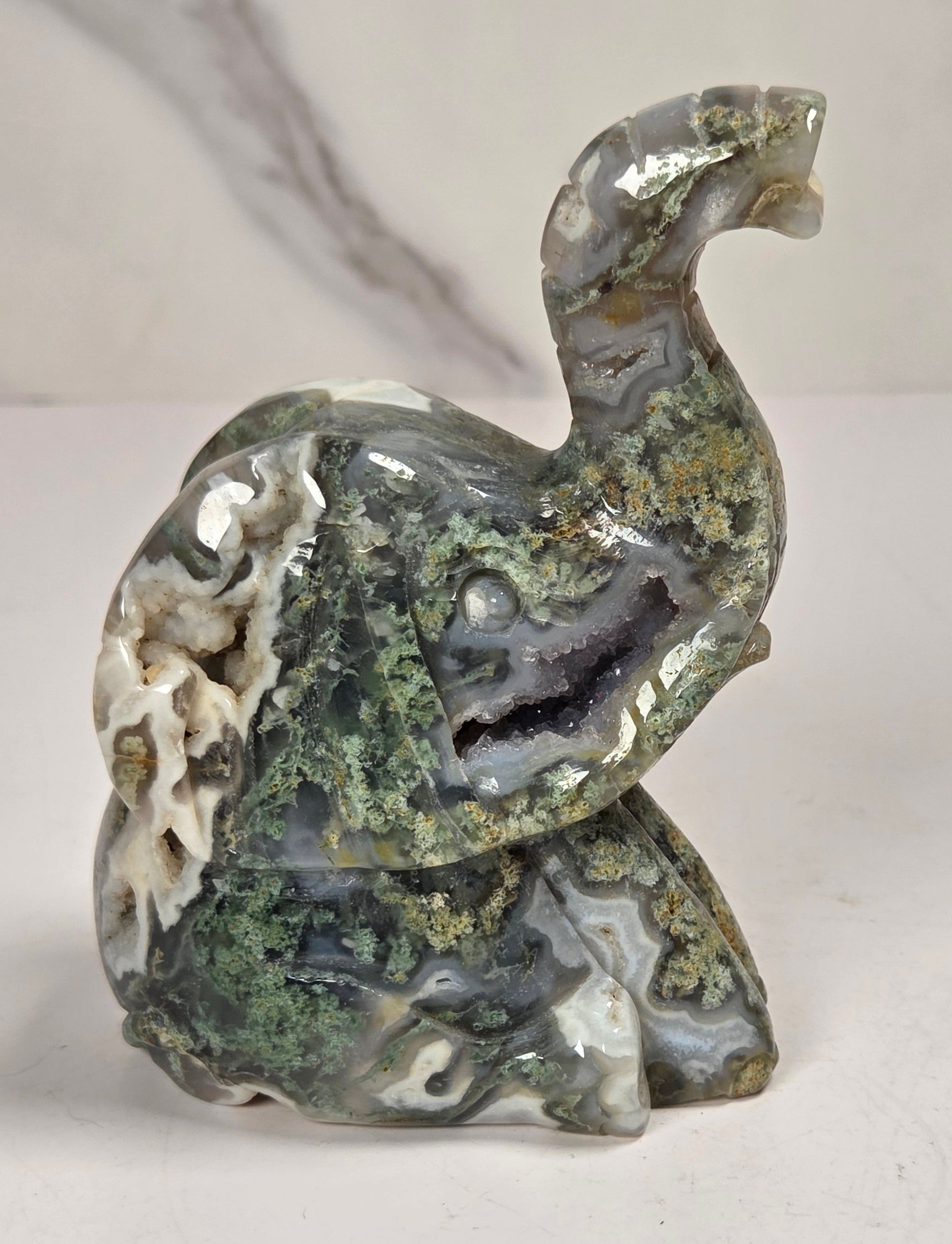 A carved elephant figurine made from polished agate stone with natural marbling in gray, white, and green, accented with brown and gold mineral inclusions, set against a light gray marble-patterned background.