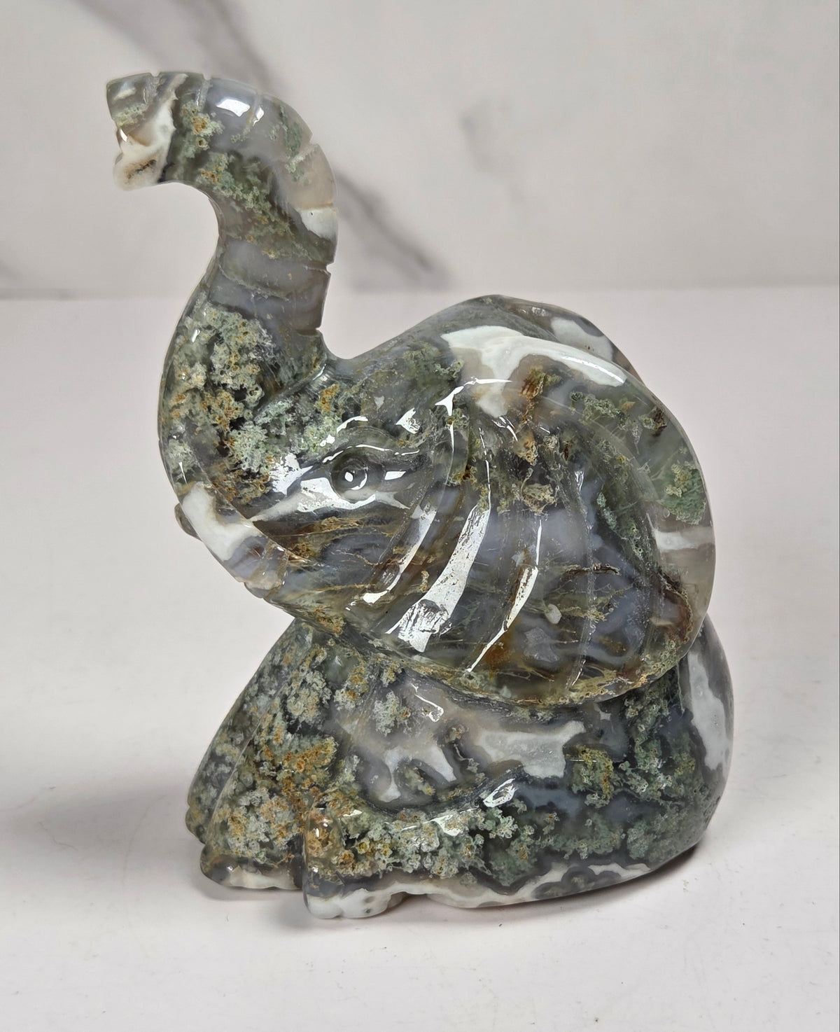 A carved elephant figurine made from polished agate stone with natural marbling in gray, white, and green, accented with brown and gold mineral inclusions, set against a light gray marble-patterned background.