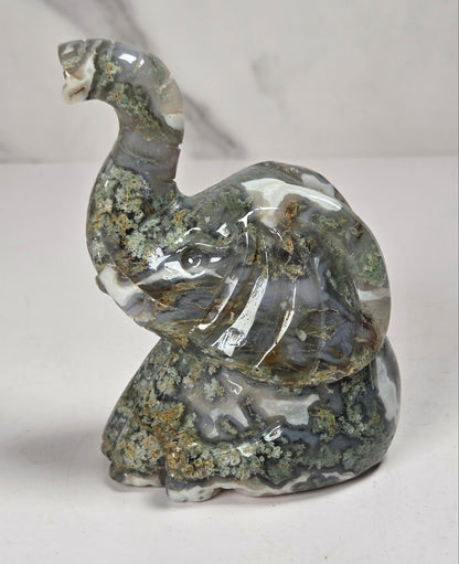 A carved elephant figurine made from polished agate stone with natural marbling in gray, white, and green, accented with brown and gold mineral inclusions, set against a light gray marble-patterned background.