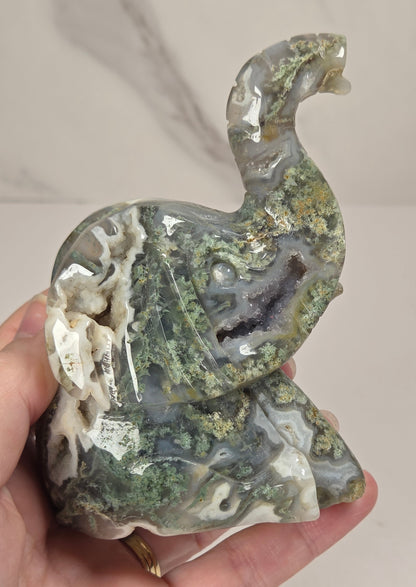A carved elephant figurine made from polished agate stone with natural marbling in gray, white, and green, accented with brown and gold mineral inclusions, set against a light gray marble-patterned background.