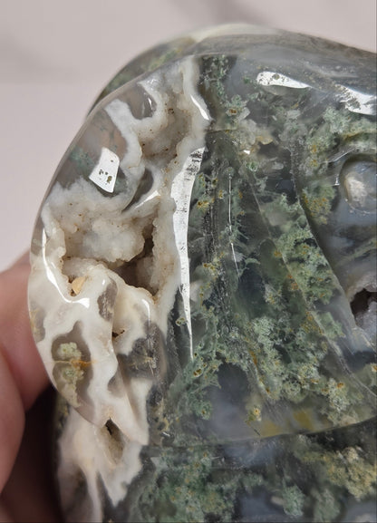 A close-up of a polished stone or mineral specimen being held in a hand. The stone has a glossy, semi-translucent surface with prominent white agate-like veining and a textured green crystalline section on the right side.