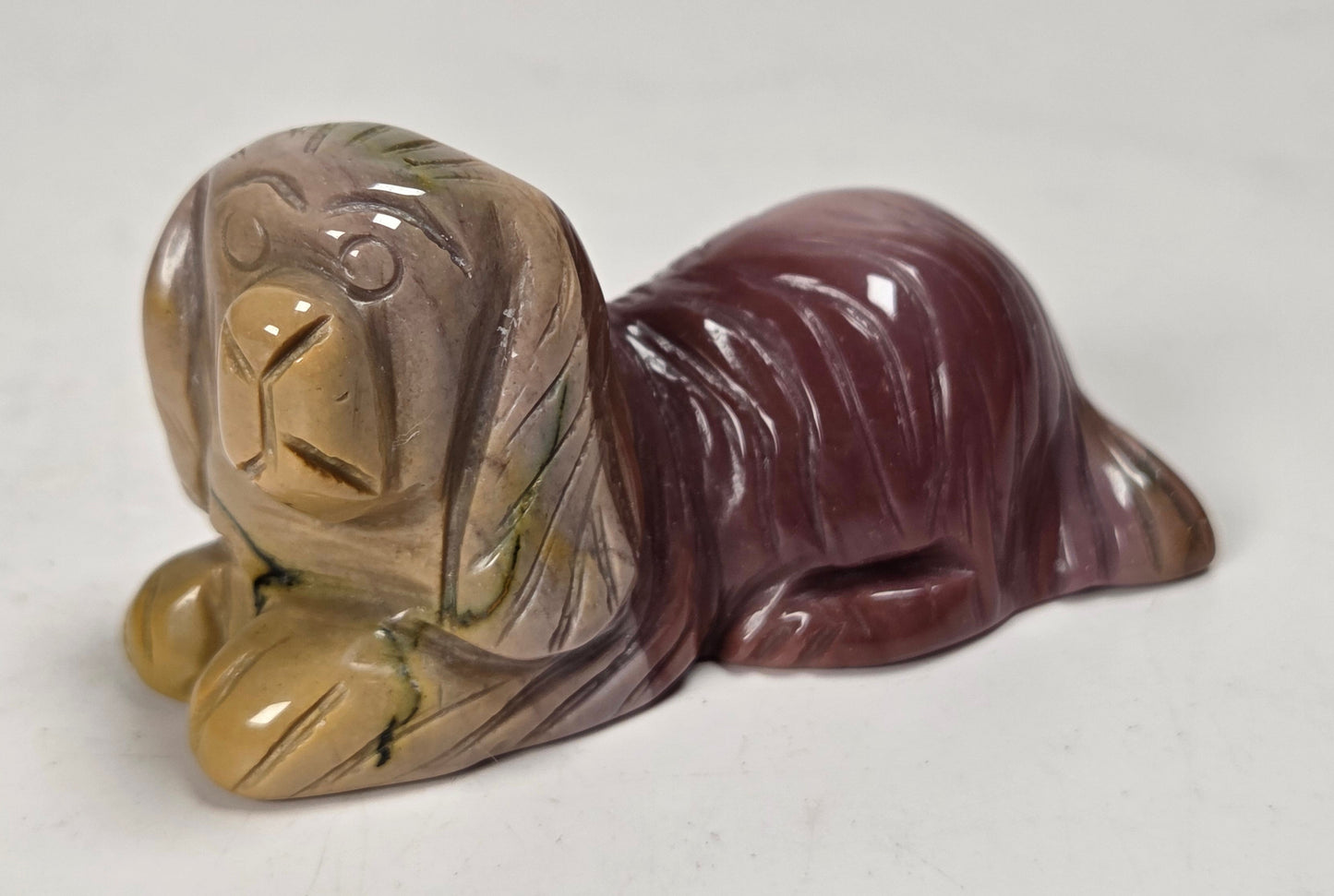 A small polished stone figurine of a lying dog, featuring a marbled coloration of tan, brown, and deep burgundy with subtle green veining, set against a plain off-white background.