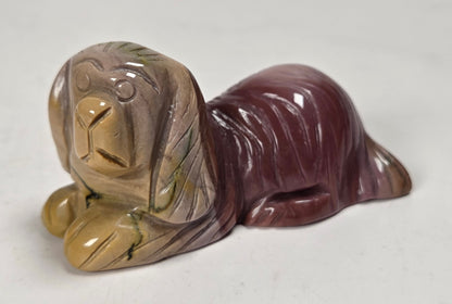 A small polished stone figurine of a lying dog, featuring a marbled coloration of tan, brown, and deep burgundy with subtle green veining, set against a plain off-white background.
