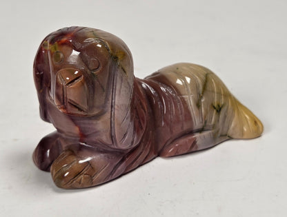 A small carved figurine of a dog lying down, made from polished stone with marbled colors of brown, purple, and yellow-green, set against a plain light gray background.