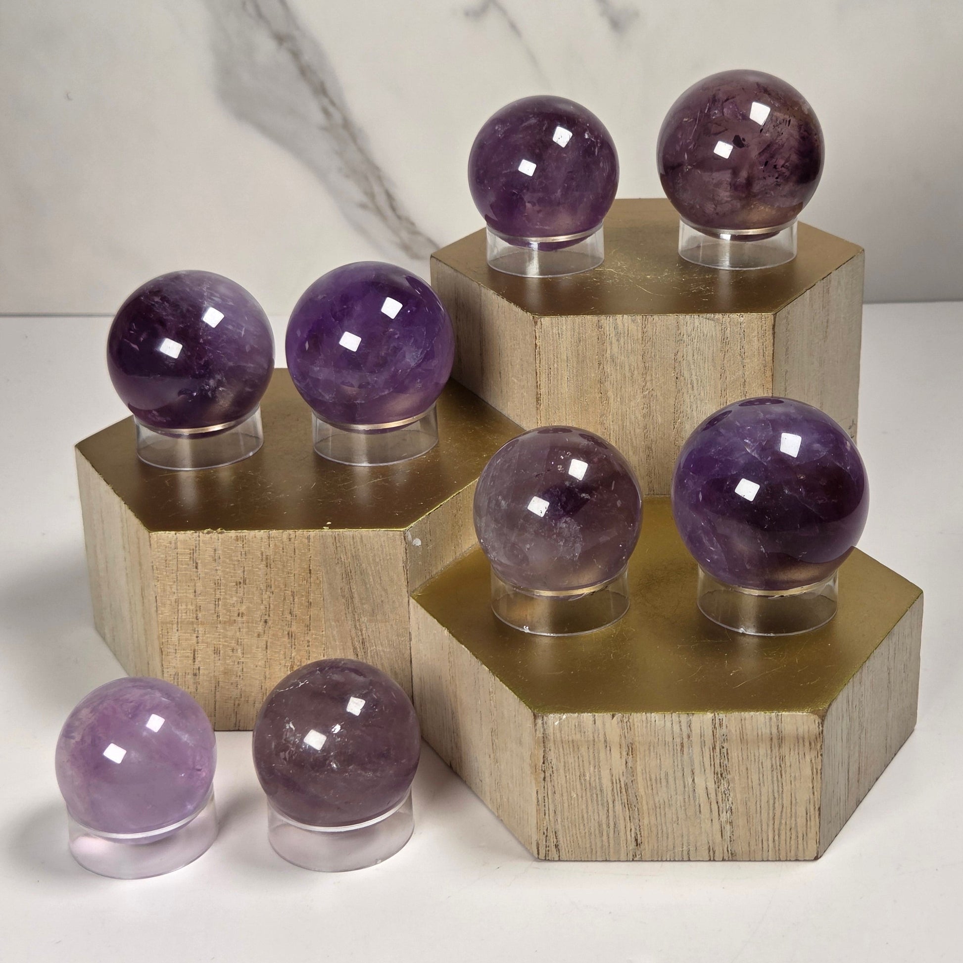 Polished purple crystal spheres of varying sizes displayed on hexagonal wooden stands against a light marble-patterned background.