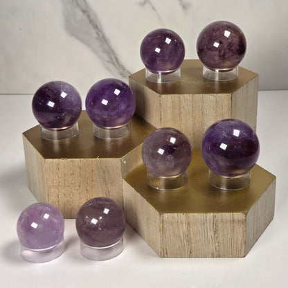 Polished purple crystal spheres of varying sizes displayed on hexagonal wooden stands against a light marble-patterned background.