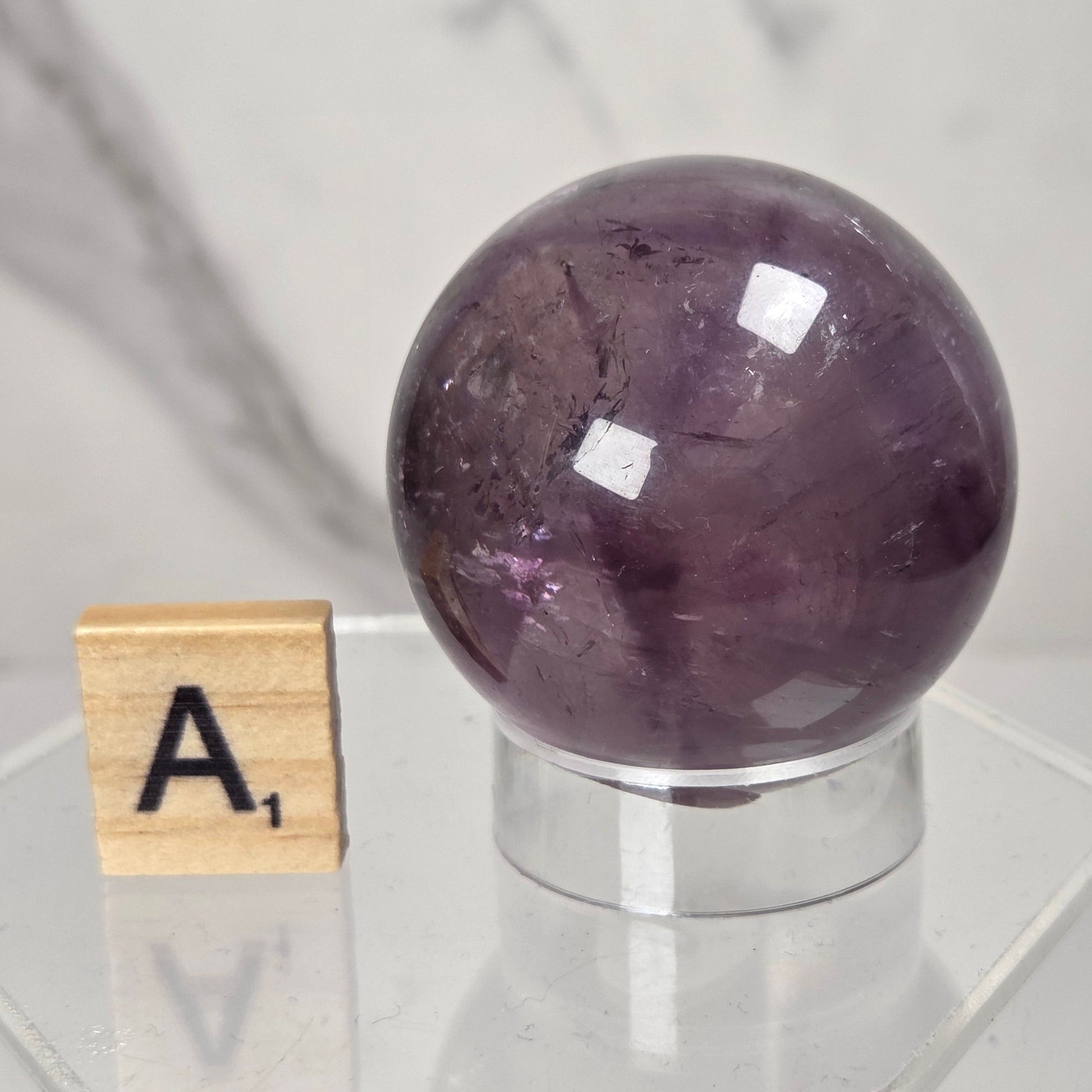 A polished purple amethyst crystal sphere displayed on a clear acrylic hexagonal pedestal next to a wooden Scrabble tile showing the letter 'A'.