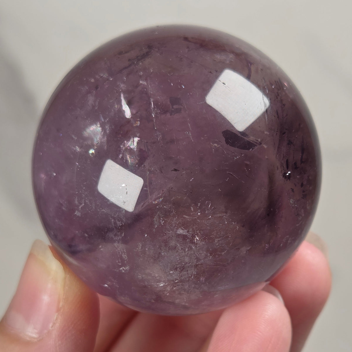 A hand holding a polished purple crystal sphere, likely amethyst, with visible natural inclusions and two white light reflections on its glossy surface.