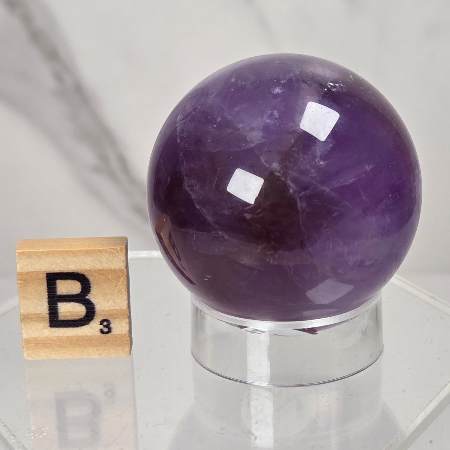 A polished purple amethyst crystal sphere displayed on a clear acrylic stand next to a wooden Scrabble tile showing the letter B, set against a light marble background.