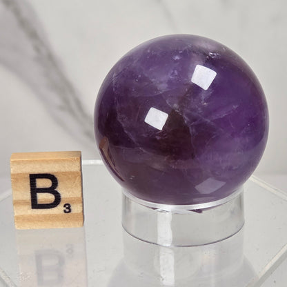 A polished purple amethyst crystal sphere displayed on a clear acrylic stand next to a wooden Scrabble tile showing the letter B, set against a light marble background.
