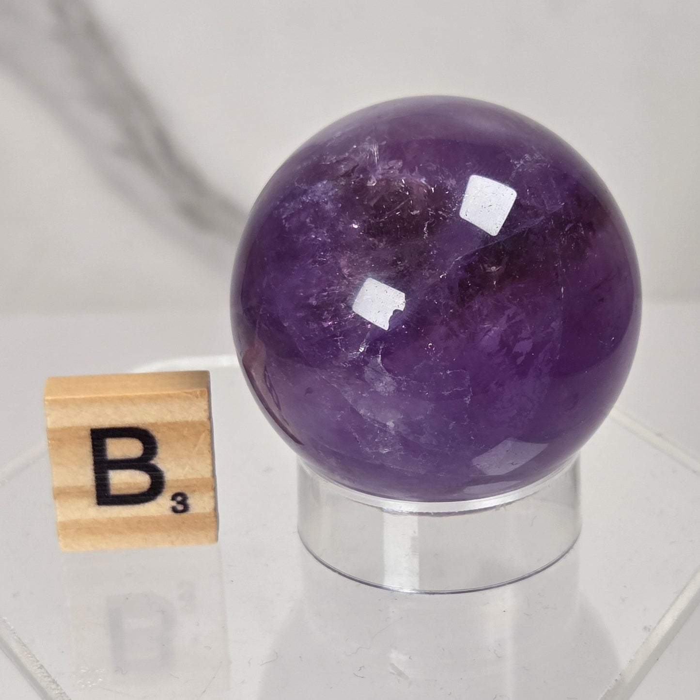 A polished purple amethyst crystal sphere displayed on a clear acrylic stand next to a wooden Scrabble tile showing the letter B, set against a light gray marble background.