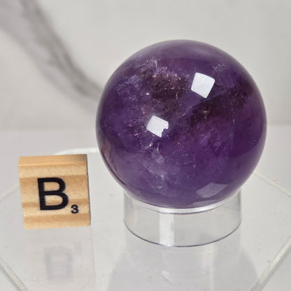 A polished purple amethyst crystal sphere displayed on a clear acrylic stand next to a wooden Scrabble tile showing the letter B, set against a light gray marble background.