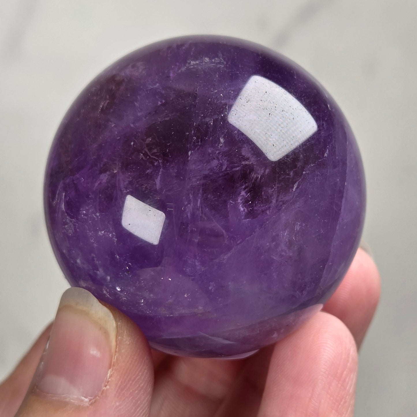 A hand holding a polished purple amethyst sphere against a light gray marble background.