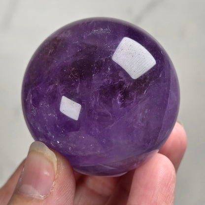A hand holding a polished purple amethyst sphere against a light gray marble background.