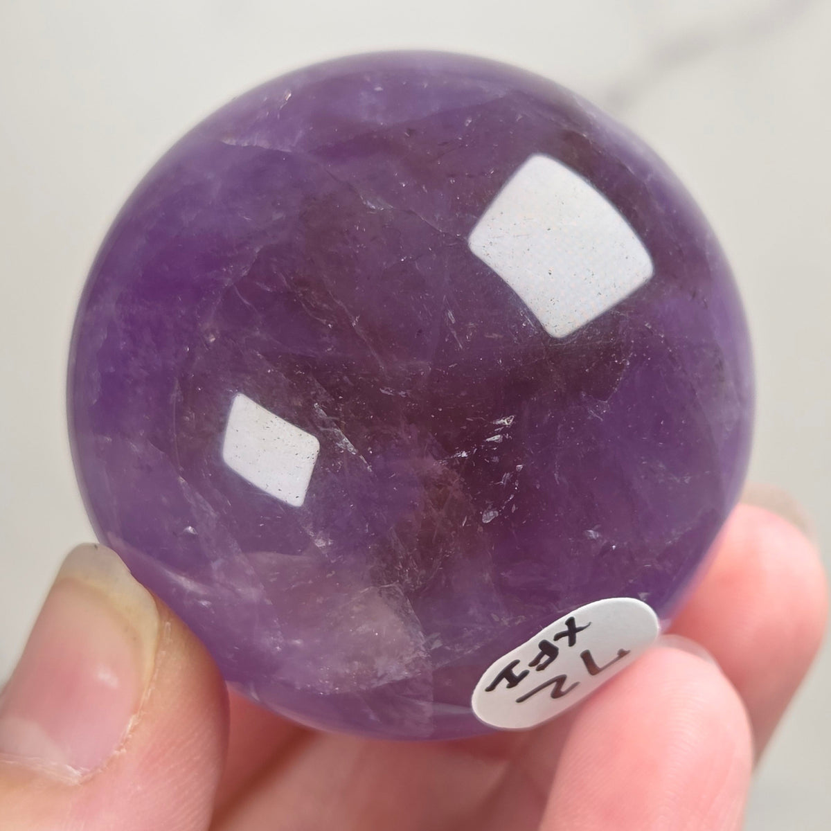 A hand holding a polished purple amethyst sphere with a small white sticker label, against a light gray marble background.