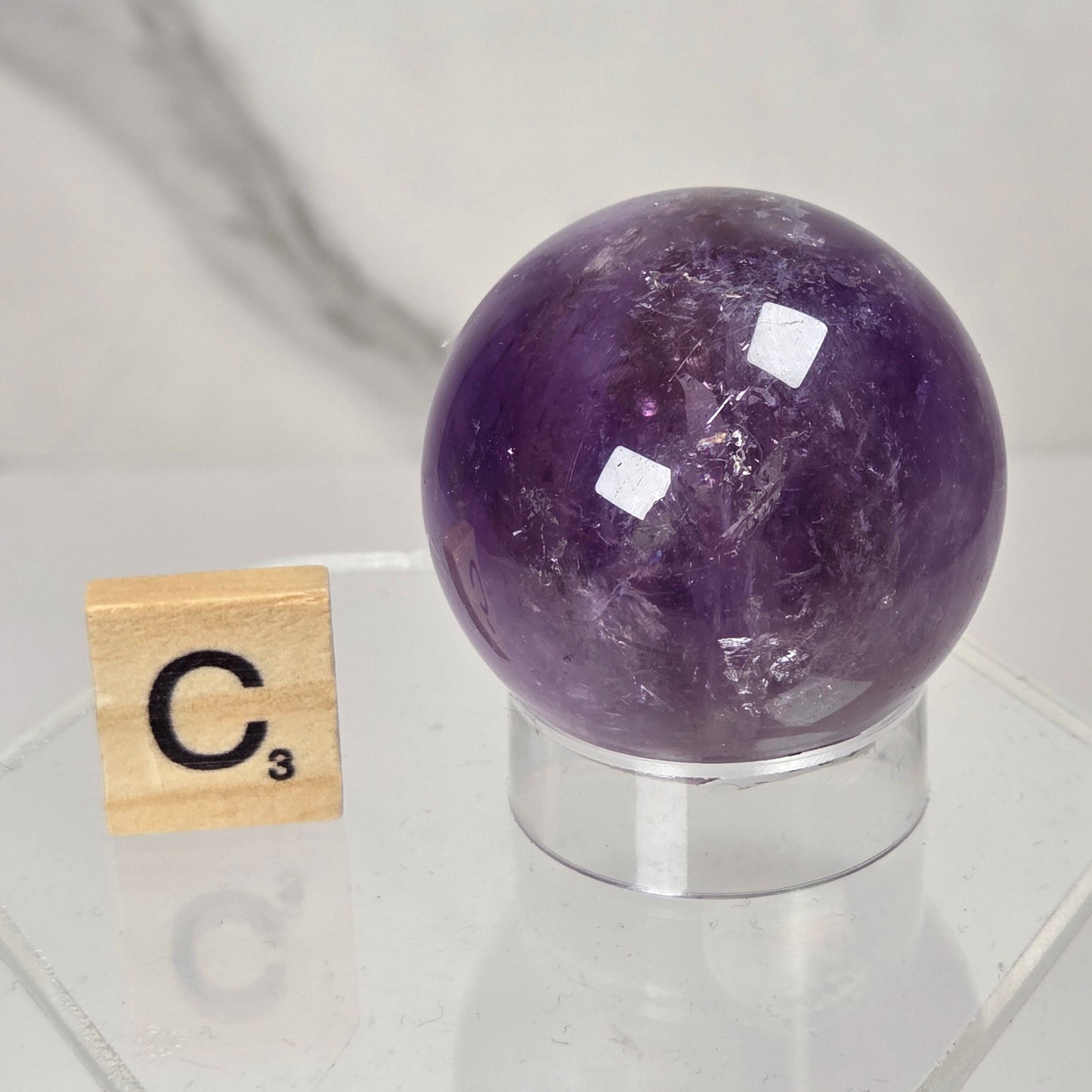 A polished purple amethyst crystal sphere on a clear acrylic stand, displayed next to a wooden Scrabble tile showing the letter C3, against a light gray marble-patterned background.