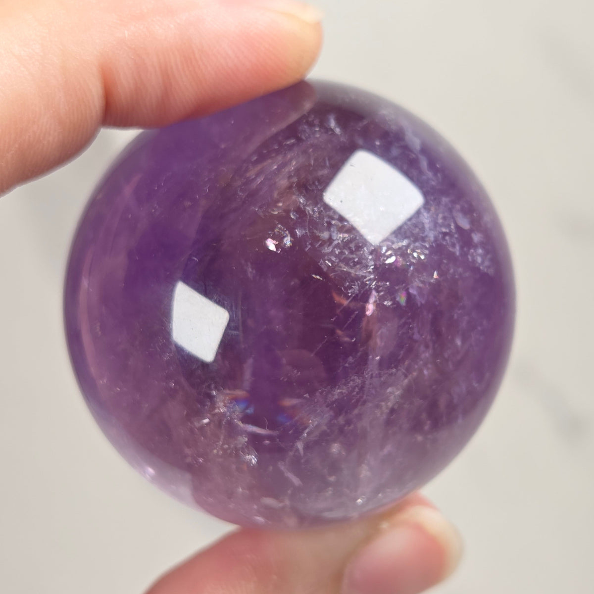 A hand holding a polished purple amethyst crystal sphere against a light gray background.